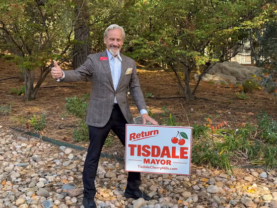 Doug Tisdale giving a thumbs-up beside his campaign sign, with autumn trees behind him
