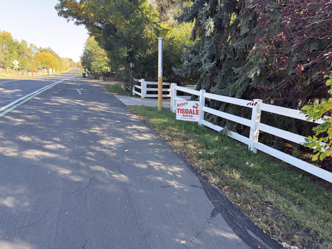A Doug Tisdale sign attached to a white fence along a tree-lined lane.